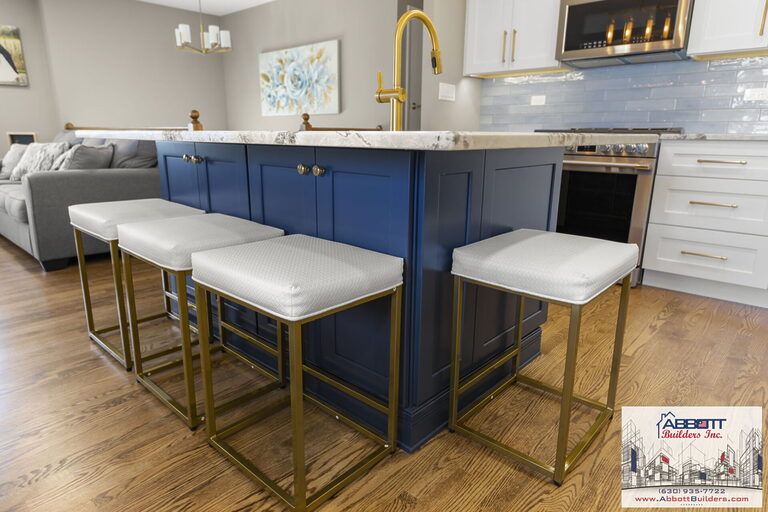 Custom kitchen island design in Lombard IL featuring navy cabinetry, marble countertop, gold faucet, and white upholstered bar stools by Abbott Builders Inc.
