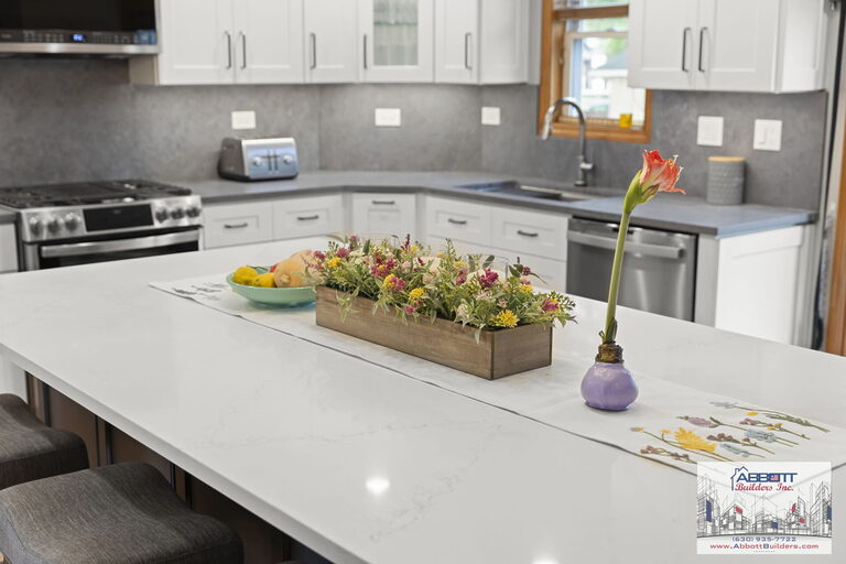 Close-up of a modern kitchen island with a white quartz countertop decorated with a wooden floral centerpiece and a single flower vase, with white shaker cabinets and gray quartz countertops in the background.
